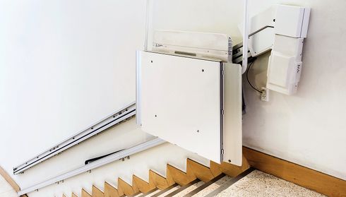 A stairlift platform attached to a staircase, featuring a white panel with railing tracks and controls for accessibility assistance.