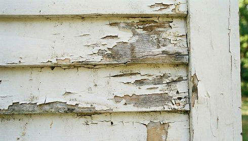 Close-up of weathered wooden siding with peeling white paint, revealing exposed wood beneath.