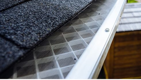 Close-up of a black asphalt shingle roof with a metal mesh gutter guard, secured by screws, preventing debris from clogging the gutter.