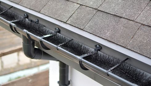 Gutter with protective brush inserts under a shingled roof, preventing debris accumulation and ensuring water flow.